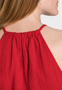 Top de tela texturizada roja con cuello redondo, detalles con volantes y un cierre de un solo botón en la parte posterior. Tirantes finos ajustables.
