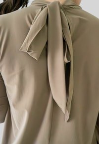Beige blouse met een grote strik op de achterkant van de hals, die een deel van de bovenrug en schouders van een persoon toont.