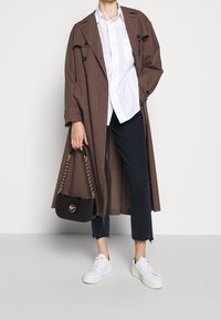 Lang brunternet frakk med stor krave, hvid skjorte under, mørke jeans med flosset kant og hvide sneakers, mens der holdes en sort kædehandtaske.