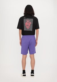Camiseta negra de manga corta con un gráfico de un corazón rojo en la parte posterior, combinada con pantalones cortos morados y zapatillas blancas con detalles en negro.