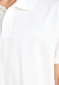 Polo blanc en tissu texturé, doté d'une patte de boutonnage à trois boutons et d'un logo en relief sur la poitrine gauche.