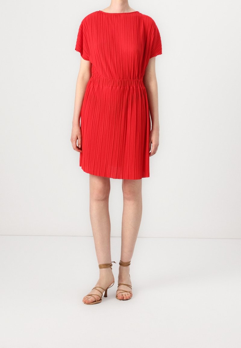 Femme portant une robe rouge plissée à manches courtes avec une taille élastique, associée à des sandales beiges à talons et à brides.