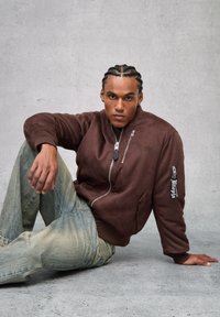 Giacca bomber marrone con una texture morbida, caratterizzata da una cerniera frontale e un logo ricamato. Abbinata a jeans di denim blu consumati.