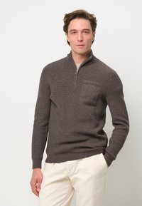 Brauner, gerippter Strickpullover mit einem Viertel-Reißverschluss und einer Brusttasche, ausgestattet mit strukturiertem Stoff und enganliegenden Bündchen. Kombiniert mit hellcremigen Hosen.