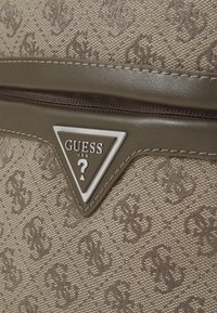 Sac en textile beige avec un motif de logo en relief, des accents lisses en vert olive, et un plaque en métal triangulaire avec le texte "GUESS".