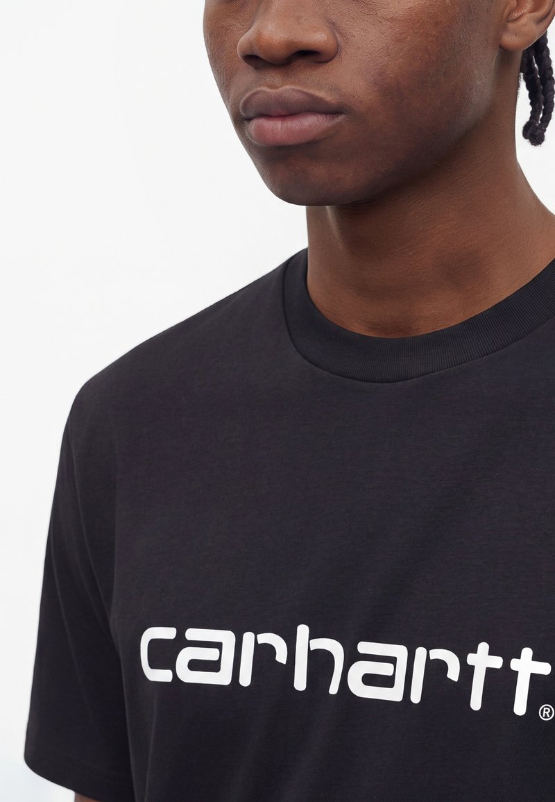 Junge Person mit dunkler Haut, die ein schwarzes T-Shirt mit weißem "carhartt"-Logo trägt, vom Nasen- bis zum Brustbereich vor einfarbigem Hintergrund.