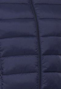 Gros plan d'une veste matelassée bleu marine avec des coutures horizontales et une fermeture éclair noire centrale.