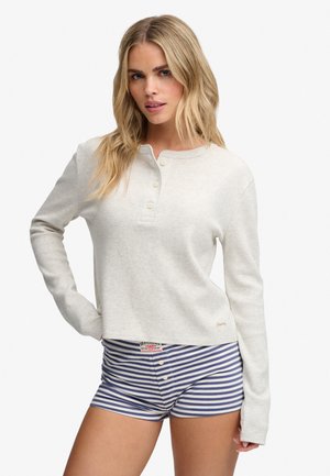 Kvinde med langt blond hår iført en lys beige langærmet henley-top og blå og hvide stribede shorts mod en hvid baggrund.