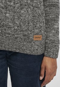 Strikkegenser i mørkegrå og hvit med et teksturert mønster, med en brun skinnlapp som viser "OXMO" ved kanten. Cord pants i marineblå.