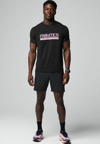 Schwarzes Fabletics-T-Shirt mit pinkem Logo, kombiniert mit schwarzen Shorts. Das Modell trägt schwarze und pinke Sportschuhe. Glatte Stofftextur.