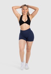 Zwarte halter sportbeha gecombineerd met marineblauwe hoge shorts. Model staat zelfverzekerd en draagt witte sportschoenen. Neutrale achtergrond.