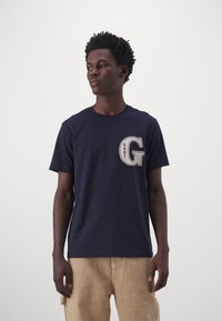 Camiseta de algodón azul marino con un gran gráfico de la letra "G" en blanco y gris en el pecho. Mangas cortas y escote redondo.