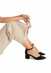 Zapatos de tacón de bloque de ante negro con correas en el tobillo. El diseño presenta una punta afilada y un acabado de tela texturizada, en contraste con pantalones beige.