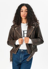 Brun skinnbikerjakke med en kortet design, sølvfarget hardware og reverskrage. Kombinert med en hvit skjorte og blå høytlivsjeans.