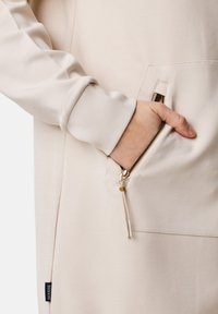Lichtbeige oversized hoodie met een ritszak, met een gladde textuur en geribde manchetten. Gouden ritshardware details zichtbaar.
