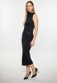 Schwarzes ärmelloses Oberteil mit einem hohen Ausschnitt, kombiniert mit einem eng anliegenden schwarzen Rock, der am Saum ausgestellt ist, ergänzt durch High-Heels.