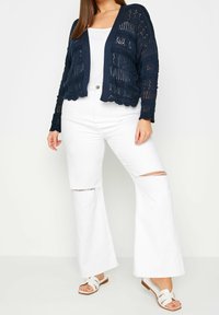 Marineblauer Strickcardigan mit offenem Muster, kombiniert mit weißen Schlagjeans mit Knieflicken und weißen Rutschsandalen.