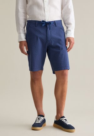 Pantalones cortos texturizados azules con cintura ajustable, que presentan bolsillos laterales y una longitud hasta medio muslo. Combinados con zapatillas navy y una camisa blanca de botones.