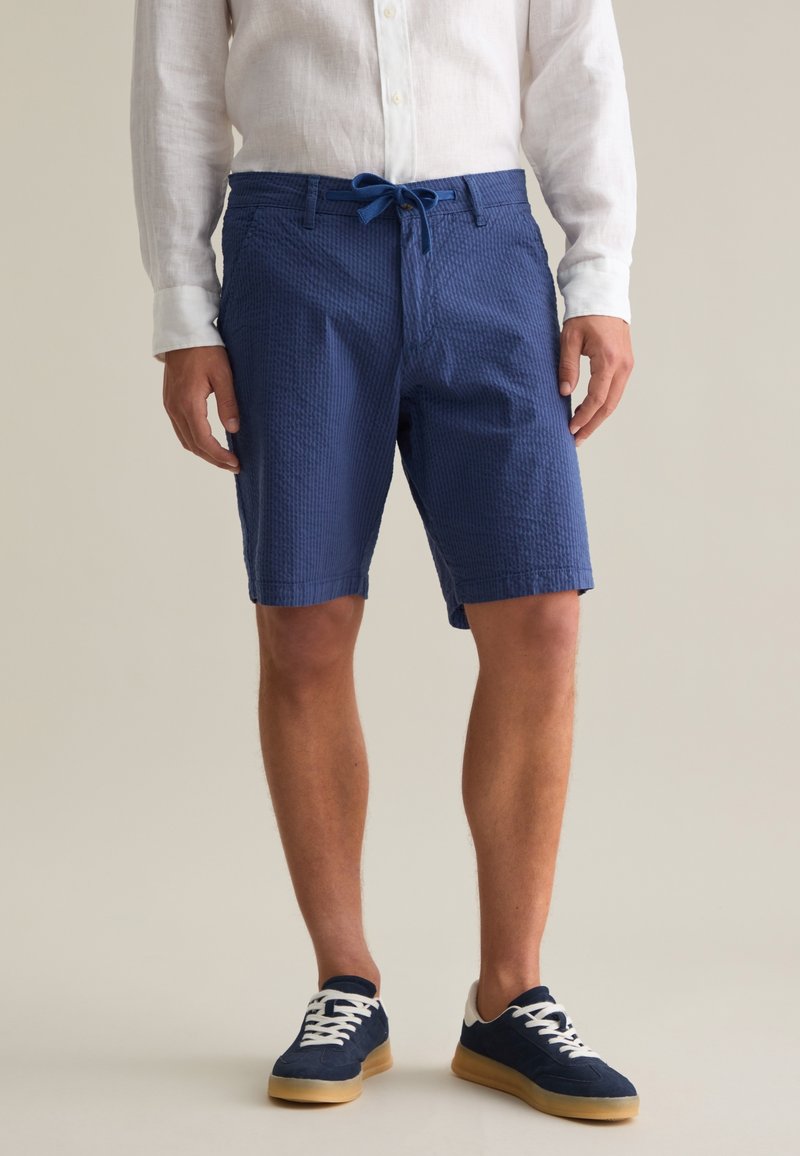 Pantalones cortos texturizados azules con cintura ajustable, que presentan bolsillos laterales y una longitud hasta medio muslo. Combinados con zapatillas navy y una camisa blanca de botones.
