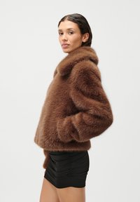 Καφέ ψεύτικη γούνα μπουφάν με βαριά υφή, που διαθέτει φαρδύ γιακά, μακριά μανίκια και κοντή γραμμή, σε συνδυασμό με μια μαύρη πλισέ φούστα.