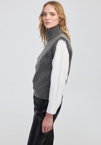 Szary, bezrękawnik o wysokim kołnierzu, wykonany z teksturowanej mieszanki wełny, noszony na białej koszuli z długim rękawem, zestawiony z czarnymi skórzanymi spodniami.