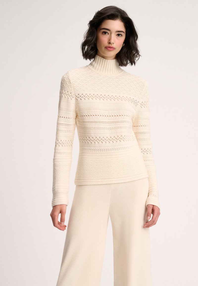 Cremfarbener, gestrickter Rollkragenpullover mit strukturierten Mustern und zarten Lochmustern, kombiniert mit passenden, leichten Hosen.