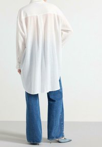Camisa blanca de manga larga con un tejido texturizado, con dobladillo curvado y un pliegue en la parte trasera, combinada con unos pantalones vaqueros de pierna ancha de color azul claro y tacones puntiagudos.