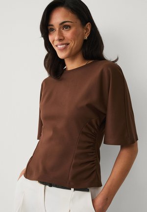 Kvinde smilende iført en brun kortærmet top med sidesamlinger og hvide bukser med hænder i lommerne mod en ensfarvet baggrund.