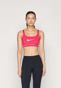 Roza sportska grudnjak s širokim naramenicama, s bijelim Nike logom i natpisom "NIKE PRO" u kontrastnom remenu. Uparen s crnim tajicama.