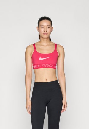 Sostenedor deportivo rosa con tirantes anchos, que presenta un logotipo blanco de Nike y el texto "NIKE PRO" en una banda contrastante. Combinado con leggings negros.