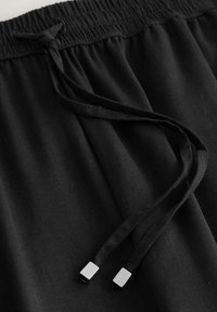 Črni elastični pas z vrvico s srebrnimi kovinskimi konci, izdelan iz mehke, gladke tkanine z rahlim sijajem.