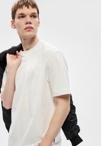 Tricou alb din bumbac cu mâneci scurte și un mic logo pe piept, purtat cu o jachetă bomber neagră aruncată pe umăr.