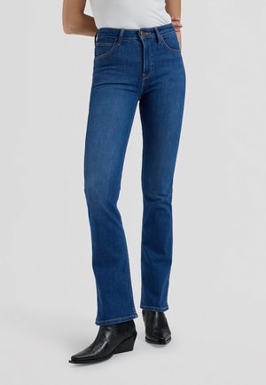 Femme portant un jean bleu taille haute droit et des bottines noires à talons sur un fond uni.