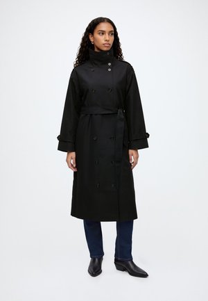 Femme aux longs cheveux bouclés portant un trench-coat noir croisé, un jean bleu foncé et des bottines noires, debout devant un fond blanc.
