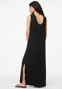Juoda maksi suknelė su iškirpta nugaros dalimi, vertikaliai raibiu tekstūru, šoninės išpjovos ir be rankovių dizainu. Dėvima su plokščiomis juodomis šlepetėmis.