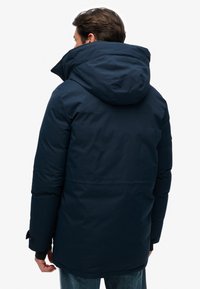 Parka isolée bleu marine avec une grande capuche, présentant une texture lisse et des poignets réglables. La vue arrière montre un design épuré et moderne.