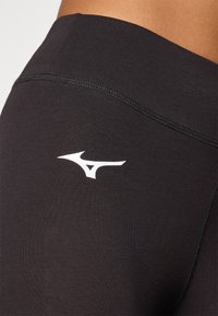Sorte atletiske leggings med en glatt, elastisk tekstur og en hvit merkevarelogo på den øvre låret. Har ren søm langs kantene.