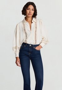 Cremefarbenes Langarmshirt mit Spitzendetails an Vorderseite und Bündchen, kombiniert mit dunklen blauen Jeans; glatter Stoff und entspannte Passform.