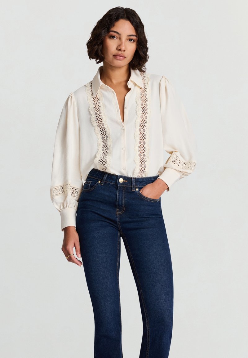 Cremefarbenes Langarmshirt mit Spitzendetails an Vorderseite und Bündchen, kombiniert mit dunklen blauen Jeans; glatter Stoff und entspannte Passform.