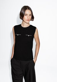 Black sleeveless top with silver accents and front pockets. Made of a smooth, fitted fabric, featuring a round neckline and clean lines.