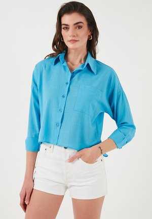Light blue button-up shirt with a collar, long sleeves rolled to the elbow, front pocket, and a relaxed fit. Paired with white shorts.