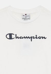 Valkoinen puuvillainen t-paita, jossa on musta "Champion"-logo kirjoitettuna kursiivilla. Sisäkauluksessa on etiketti, jossa on koko XS ja hoito-ohjeet.