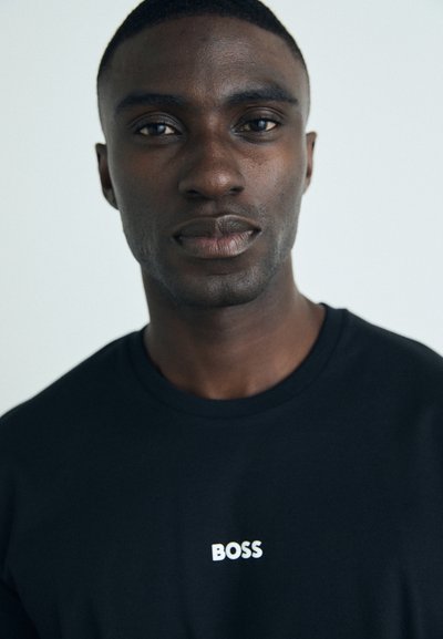 T-shirt en coton noir avec un logo "BOSS" blanc sur la poitrine. Texture douce avec un col rond. Visage du modèle partiellement visible.
