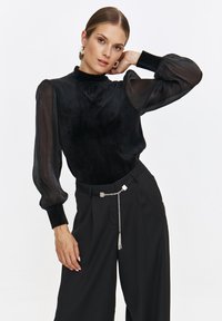 Czarna aksamitna bluzka z długimi przezroczystymi rękawami i wysokim kołnierzem, zestawiona z czarnymi spodniami o wysokim stanie i metalicznym paskiem łańcuchowym.