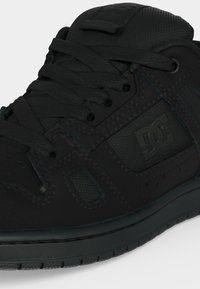 Gros plan sur une chaussure de skate DC noire montrant les lacets, les détails perforés, les panneaux en mesh et le logo DC embossé sur le côté.