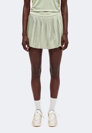 Personne portant une jupe plissée vert pâle avec le texte "ENDLESS", des chaussettes blanches et des baskets blanches, se tenant devant un fond uni.
