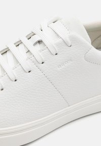 Baskets blanches en cuir avec une surface texturée, bout rond et design à lacets blancs. Présente un logo discret sur le côté et une semelle en caoutchouc.