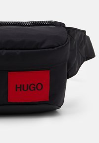 Pochette en tissu noir avec une finition texturée, dotée d'un patch rectangulaire rouge avec "HUGO" en lettres noires et d'une fermeture éclair noire.