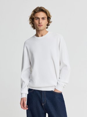 Giovane uomo con capelli castano chiaro e ricci, che indossa un maglione bianco semplice a maniche lunghe e jeans blu scuro, in piedi contro uno sfondo grigio.
