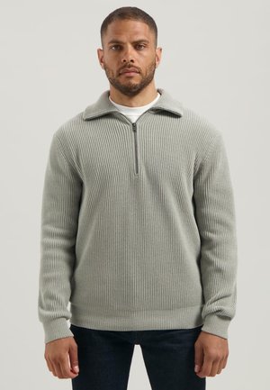 Man in een lichtgrijs geribbeld gebreid zip-up sweater met donkere broek, staande tegen een effen lichte achtergrond.